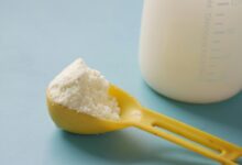 A yellow spoon filled with powdered milk, next to a measuring bottle, on a blue background.