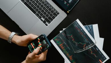 Hands using smartphone beside laptop with stock charts, showcasing digital trading.