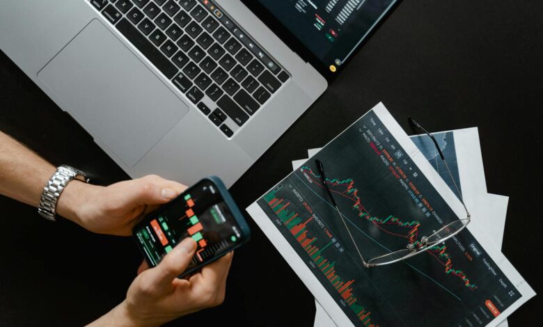 Hands using smartphone beside laptop with stock charts, showcasing digital trading.